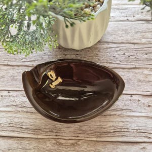 Könnte beinhalten: Eine dunkelbraune, ovale Keramikschale mit einer glatten, glänzenden Oberfläche. Die Schale verfügt über ein kleines, goldfarbenes Emblem in Form einer Harfe mit einem Schriftband, das in der Mitte der Innenfläche angebracht ist.