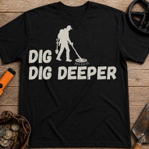 Op de afbeelding: Een zwart t-shirt met korte mouwen en een beige afbeelding van een persoon die een metaaldetector gebruikt en een schep draagt. De tekst "DIG DIG DEEPER" is in een beige, verweerd lettertype onder de illustratie van de zoeker gedrukt.