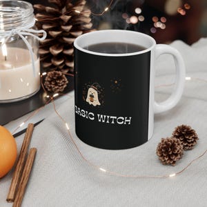 May include: A white ceramic mug with a black exterior featuring a graphic of a small ghost wearing a witch hat and holding a coffee cup. The text "BASIC WITCH" is printed in white below the ghost, surrounded by small orange star accents.