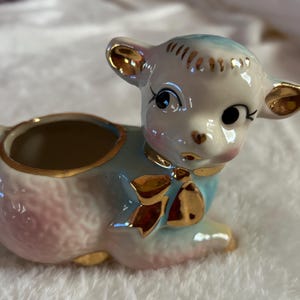 May include: A ceramic planter shaped like a lamb with a pink, blue, and white glaze. The lamb has gold accents on its face and a gold bow around its neck.