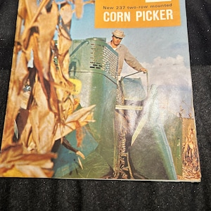 May include: Vintage advertisement for a John Deere New 237 two-row mounted corn picker. The image features a man standing near the green and yellow machinery, with a blue sky background and dried corn stalks.