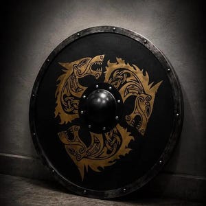 May include: A circular shield featuring a matte black surface with a metallic gold design of three snarling wolf heads arranged in a triangular pattern. The shield has a dark metal rim with visible rivets and a central black metal dome or boss.