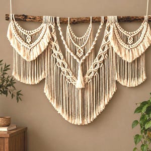 Può includere: Un grande arazzo da parete in macramè color bianco sporco appeso a un ramo di legno scuro. L'oggetto presenta complessi motivi a nodi, sezioni di frange sovrapposte e una nappa centrale, creando un design simmetrico.