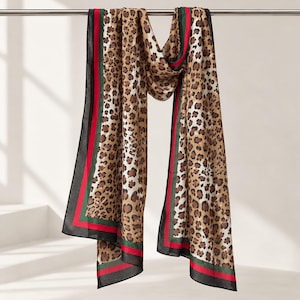 May include: A rectangular scarf featuring a brown and black leopard print pattern. The scarf is bordered by a thin green stripe and a wider red stripe, finished with a solid black edge along the perimeter. The fabric appears lightweight and smooth.