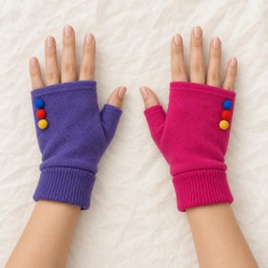 May include: A pair of fingerless gloves featuring a ribbed cuff and three small decorative pom-poms on the back of each hand. One glove is purple and the other is bright pink, both made of a soft, solid-colored fabric material.