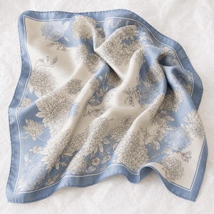 Op de afbeelding: Een vierkante sjaal met een stoffige blauwe rand en een crèmekleurig midden. De stof is versierd met een gedetailleerd bloemenpatroon van chrysanten in tinten blauw en donkergrijze lijntekeningen voor een zachte en elegante uitstraling.