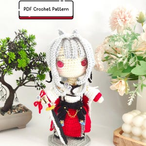 May include: A crochet doll with white hair, red eyes, and a pale complexion, wearing a white, black, and red outfit with a sword accessory. The doll stands on a gray base and includes a label that reads PDF Crochet Pattern in black text.