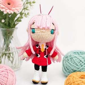 May include: A handcrafted crochet doll featuring long pink hair, two red antennae, and large teal eyes. The doll wears a red jacket with black accents, a yellow tie, black leggings, and white boots with black soles, all constructed from yarn.