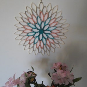 May include: A white wall with a large paper flower made from rolled paper tubes. The flower is in shades of white, gray, pink, and teal. There are pink flowers in the bottom of the image.
