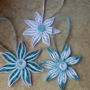 May include: Three paper quilled ornaments in the shape of flowers. Two of the flowers are white with a teal center and one flower is teal with a white center.