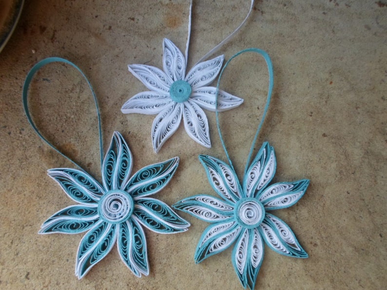 May include: Three paper quilled ornaments in the shape of flowers. Two of the flowers are white with a teal center and one flower is teal with a white center.