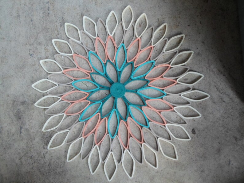 May include: A paper flower wall decor made from rolled paper, featuring a circular design with overlapping petals in shades of white, peach, teal, and blue.