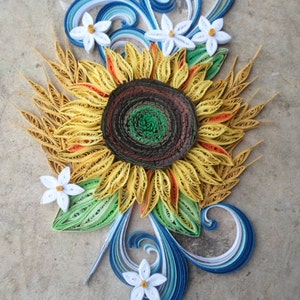 Puede incluir: Una obra de arte de papel quilling que representa un girasol con p&eacute;talos amarillos, un centro marr&oacute;n y hojas verdes. El girasol est&aacute; rodeado de flores blancas y remolinos azules y blancos.