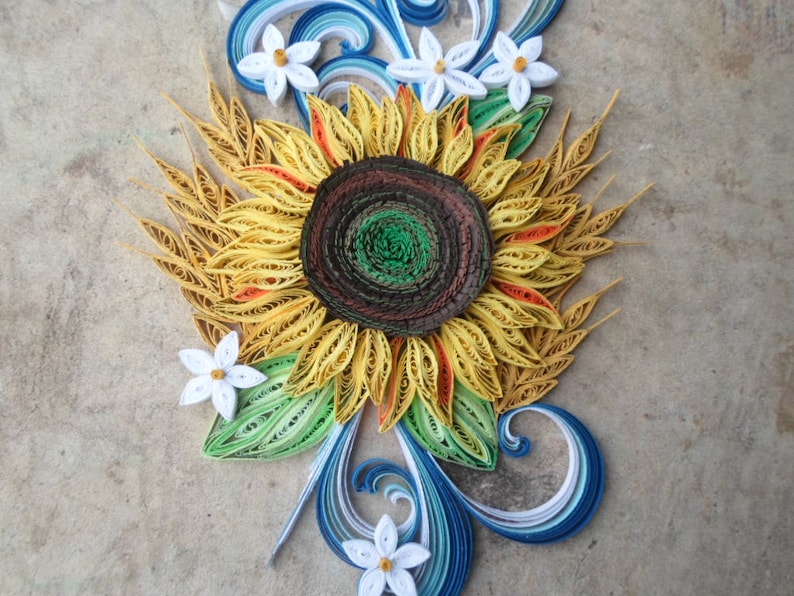 Puede incluir: Una obra de arte de papel quilling que representa un girasol con p&eacute;talos amarillos, un centro marr&oacute;n y hojas verdes. El girasol est&aacute; rodeado de flores blancas y remolinos azules y blancos.