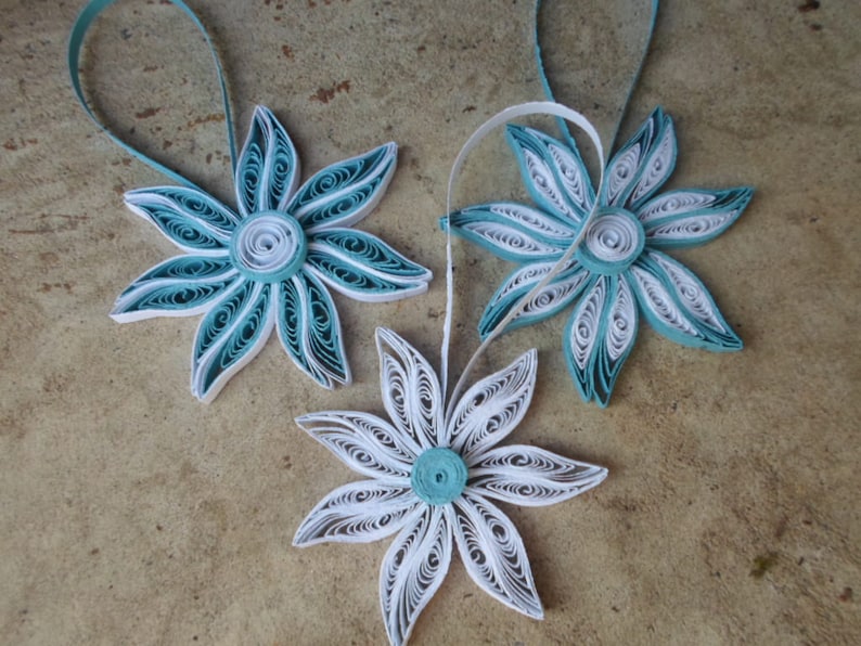 May include: Three paper quilled ornaments in the shape of flowers. Two are teal blue with white centers and one is white with a teal blue center.