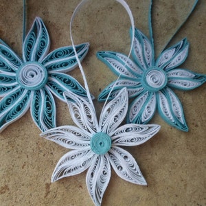 May include: Three paper quilled ornaments in the shape of flowers. Two are teal blue and white, and one is white. They are hanging from a string.