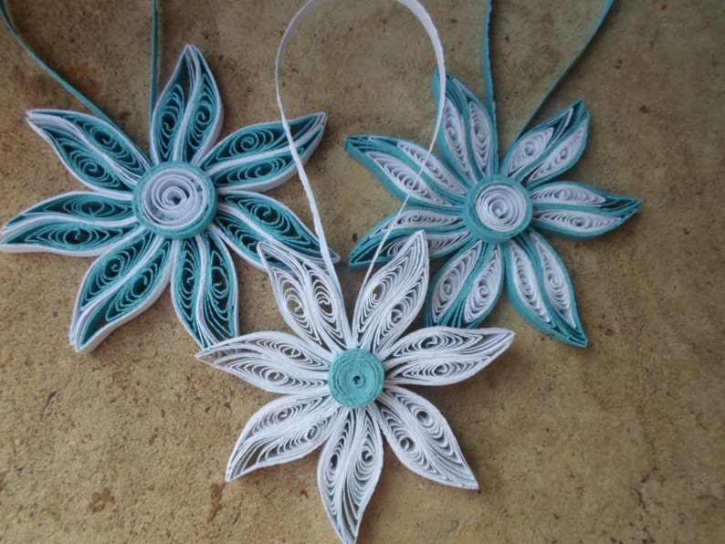 May include: Three paper quilled ornaments in the shape of flowers. Two are teal blue and white, and one is white. They are hanging from a string.