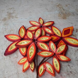 May include: A cluster of paper quilled flowers in shades of red, orange, and yellow. The flowers are arranged in a bouquet-like shape and have a delicate, intricate design.