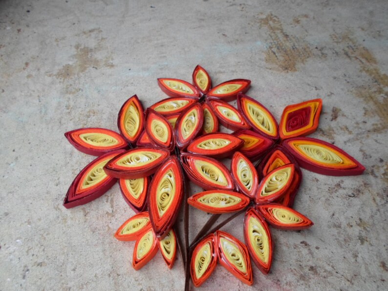 May include: A cluster of paper quilled flowers in shades of red, orange, and yellow. The flowers are arranged in a bouquet-like shape and have a delicate, intricate design.