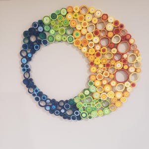 May include: A colorful, circular wall art piece. The artwork is composed of rolled paper in a gradient of colors, transitioning from blue to green, yellow, and orange. The design is mounted on a white wall, creating a vibrant and eye-catching display.