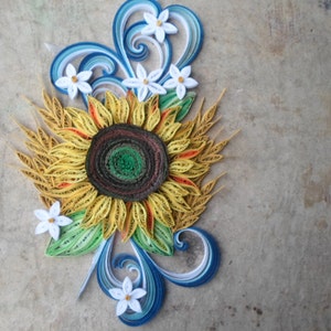 Puede incluir: Una obra de arte de quilling de papel que representa un girasol con p&eacute;talos amarillos, un centro marr&oacute;n y hojas verdes. El girasol est&aacute; rodeado de flores blancas y remolinos azules.
