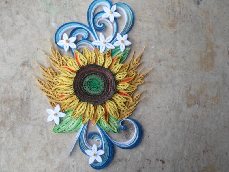 Puede incluir: Una obra de arte de quilling de papel que representa un girasol con p&eacute;talos amarillos, un centro marr&oacute;n y hojas verdes. El girasol est&aacute; rodeado de flores blancas y remolinos azules.