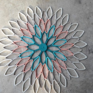 May include: A large paper flower wall decor made from rolled paper. The flower is made up of multiple layers of paper in shades of white, peach, teal, and green.