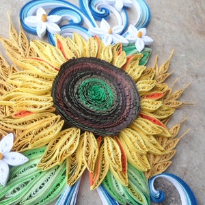 Puede incluir: Una obra de arte de quilling de papel que representa un girasol con p&eacute;talos amarillos, un centro marr&oacute;n y hojas verdes. Flores de papel blanco est&aacute;n dispuestas alrededor del girasol.