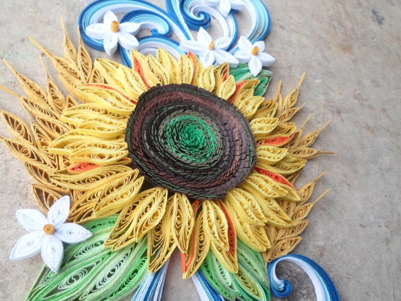 Puede incluir: Una obra de arte de quilling de papel que representa un girasol con p&eacute;talos amarillos, un centro marr&oacute;n y hojas verdes. Flores de papel blanco est&aacute;n dispuestas alrededor del girasol.
