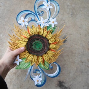 Puede incluir: Un girasol de papel quilling con un centro marr&oacute;n y p&eacute;talos amarillos. El girasol est&aacute; rodeado de flores blancas de papel quilling y tallos de trigo. El fondo es una superficie de hormig&oacute;n gris claro.