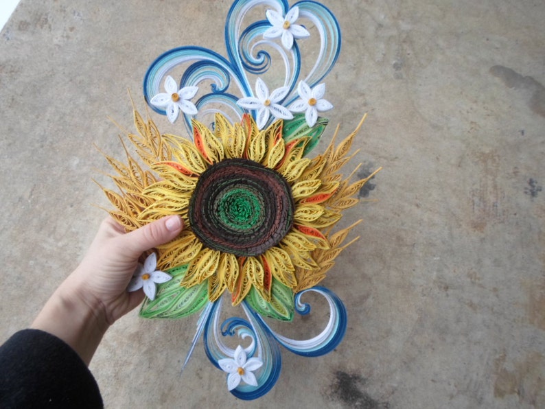 Puede incluir: Un girasol de papel quilling con un centro marr&oacute;n y p&eacute;talos amarillos. El girasol est&aacute; rodeado de flores blancas de papel quilling y tallos de trigo. El fondo es una superficie de hormig&oacute;n gris claro.