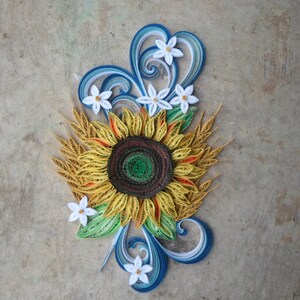 Puede incluir: Una obra de arte de quilling de papel que presenta un girasol amarillo con un centro marr&oacute;n rodeado de flores blancas y remolinos azules y blancos.
