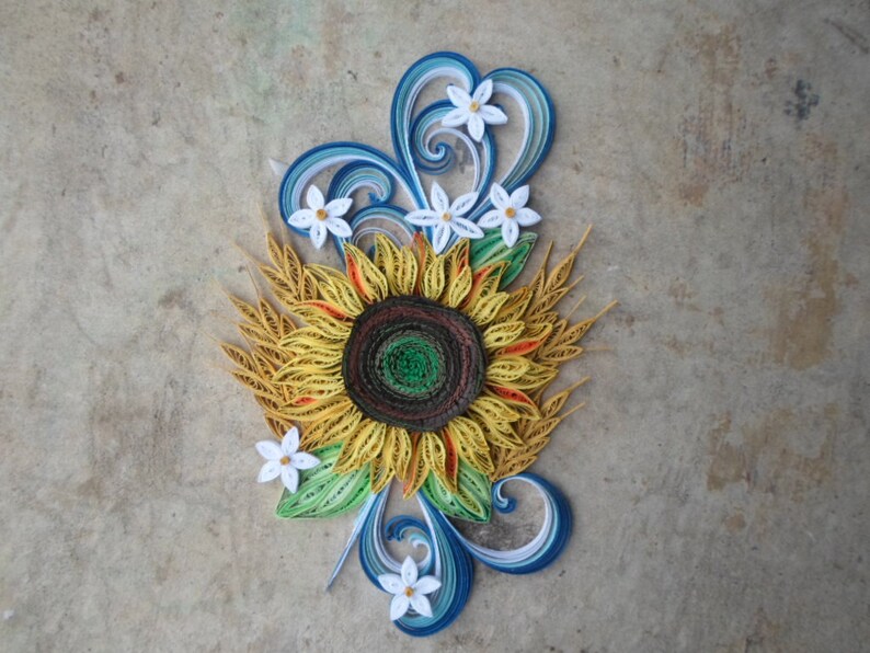 Puede incluir: Una obra de arte de quilling de papel que presenta un girasol amarillo con un centro marr&oacute;n rodeado de flores blancas y remolinos azules y blancos.