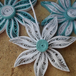 May include: Three paper flowers, one white and two teal blue, with a layered, curled design. The flowers are made of paper and have a delicate, intricate look.