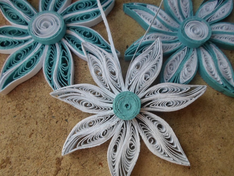 May include: Three paper flowers, one white and two teal blue, with a layered, curled design. The flowers are made of paper and have a delicate, intricate look.
