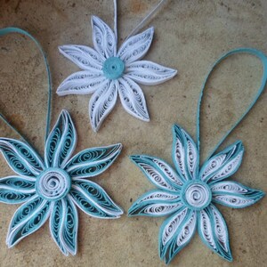 May include: Three paper quilled ornaments in the shape of flowers. Two of the ornaments are white with a teal center and one is teal with a white center.