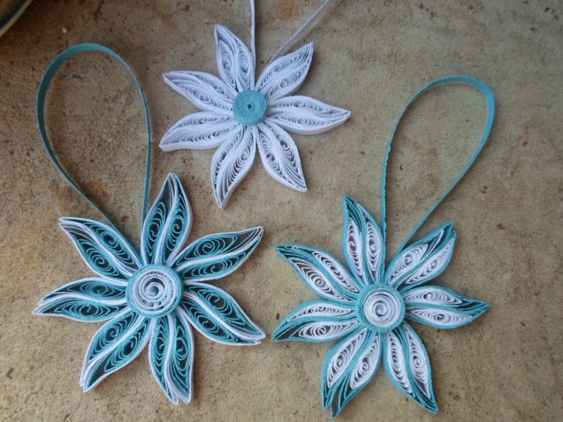 May include: Three paper quilled ornaments in the shape of flowers. Two of the ornaments are white with a teal center and one is teal with a white center.