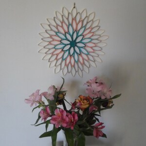 May include: A white wall with a teal, pink, and white paper flower wall hanging. Below the wall hanging is a bouquet of pink and orange flowers in a clear glass vase.