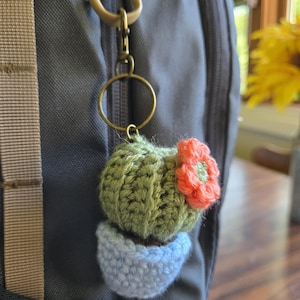 May include: A crocheted cactus keychain featuring a light green textured cactus body with a small orange flower attached to the side, sitting in a light blue crocheted pot, suspended from a bronze-colored metal ring and a lobster claw clasp.