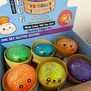 May include: A display box containing six round, squishy bun toys in various colors including green, blue, orange, yellow, and purple. Each toy features a smiling face and glittery texture, housed in individual tan plastic containers resembling bamboo steamers.