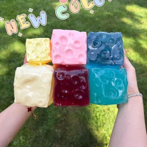 May include: Six cube-shaped objects with circular indentations resembling Swiss cheese. The colors include pale yellow, light pink, dark blue, cream, deep red, and bright turquoise. The text NEW COLOR is displayed in a playful, outlined font.