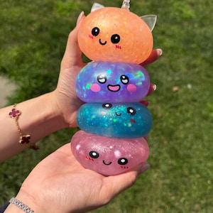 May include: A stack of four translucent, squishy, gel-like spheres with glittery inclusions. Each sphere features a printed cartoon face with large black eyes, a small mouth, and pink cheeks. The top sphere has small cat ears and a tiny horn.