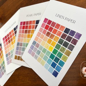 May include: Three white paper sample cards labeled Linen Paper, Pearl Paper, and Cotton Paper, each featuring a grid of small, hand-painted watercolor squares in a spectrum of colors including red, orange, yellow, green, blue, and purple.