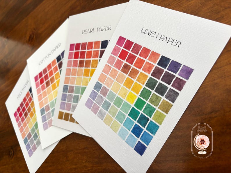 May include: Three white paper sample cards labeled Linen Paper, Pearl Paper, and Cotton Paper, each featuring a grid of small, hand-painted watercolor squares in a spectrum of colors including red, orange, yellow, green, blue, and purple.