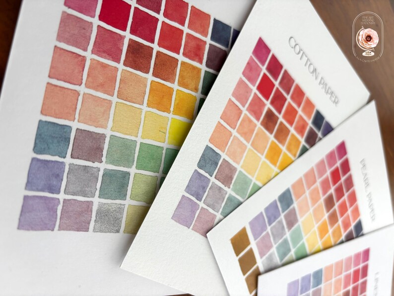 May include: Three white paper sheets featuring grids of watercolor swatches in a spectrum of colors including red, orange, yellow, green, blue, and purple. The sheets are labeled with the text COTTON PAPER and PEARL PAPER in black ink.