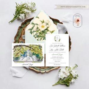 May include: A wedding invitation set featuring a white card with black text and a watercolor illustration of a fountain in Savannah, Georgia. An envelope with a floral interior lining is included, accompanied by white flowers and a logo.