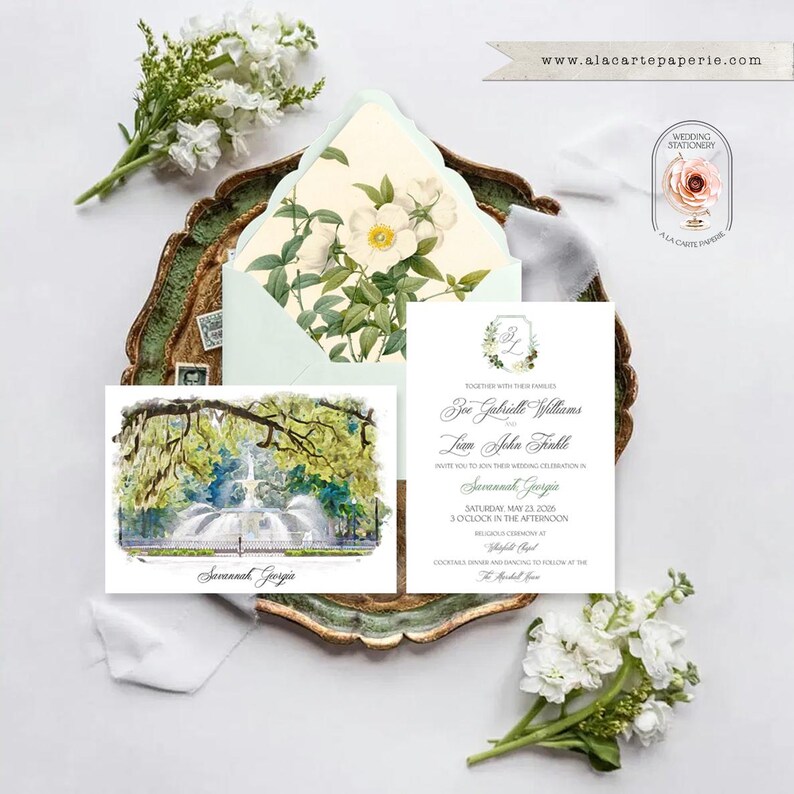 May include: A wedding invitation set featuring a white card with black text and a watercolor illustration of a fountain in Savannah, Georgia. An envelope with a floral interior lining is included, accompanied by white flowers and a logo.