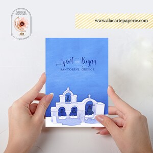 Pode incluir: Um convite de casamento em aquarela azul com letras brancas que diz "April e Bryan Santorini, Gr&eacute;cia". O convite apresenta uma silhueta branca de uma igreja grega com sinos azuis.