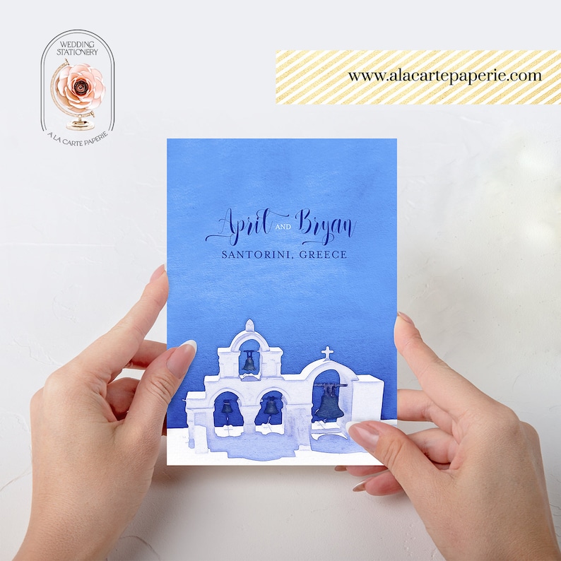 Pode incluir: Um convite de casamento em aquarela azul com letras brancas que diz "April e Bryan Santorini, Gr&eacute;cia". O convite apresenta uma silhueta branca de uma igreja grega com sinos azuis.