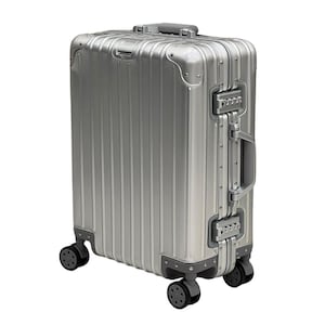 May include: A silver metallic suitcase with a vertical ribbed exterior design. The luggage features reinforced corner caps, a retractable top handle, a side handle, and two integrated combination locks. It stands on four black multi-directional wheels.